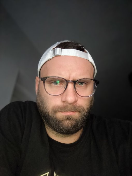 A close-up selfie of a bearded man wearing glasses and a backward white cap against a dark background.