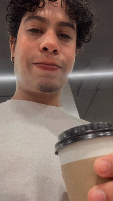 A selfie of a young man with curly hair wearing a light grey t-shirt, holding a takeaway coffee cup.