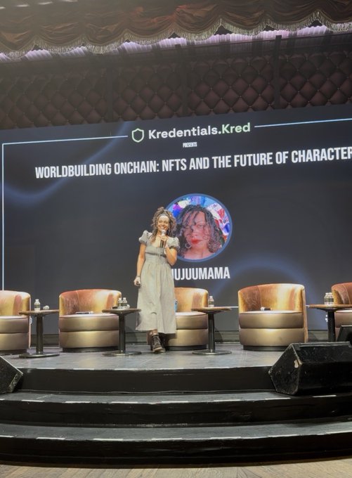 A woman on stage speaking at a panel titled 'WORLDBUILDING ONCHAIN: NFTS AND THE FUTURE OF CHARACTER' presented by Kredentials.Kred, with her avatar 'JUJUUMAMA' displayed on the screen.