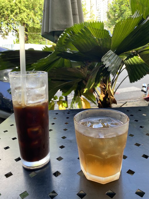 Two iced drinks, one dark (likely cola) in a tall glass with a straw and one lighter amber drink in a shorter glass, sit on an outdoor table with palm fronds in the background.