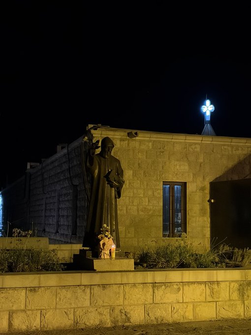 A nighttime photo of a stone building with a glowing cross on top, featuring a statue of a religious figure holding a staff.