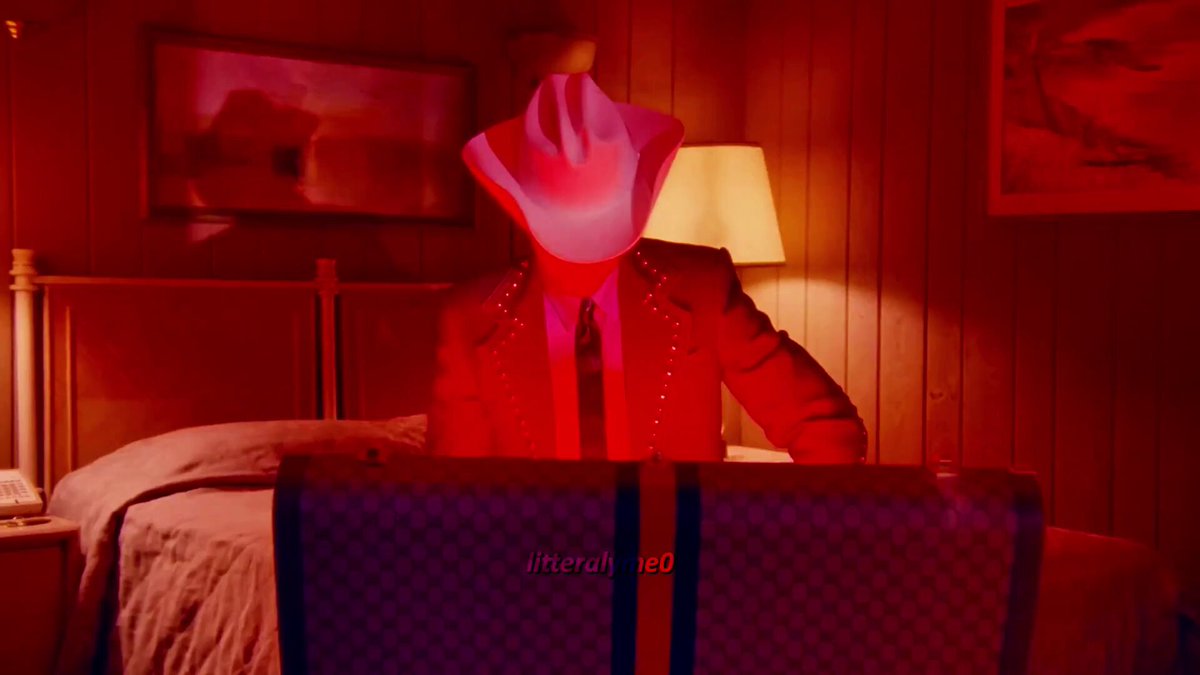 The image shows a man wearing a cowboy hat sitting at a desk with his laptop open, possibly working or browsing the internet. The room has red walls and a bed visible in the background. There are also two chairs present in the scene - one near the man's chair and another further away from him.