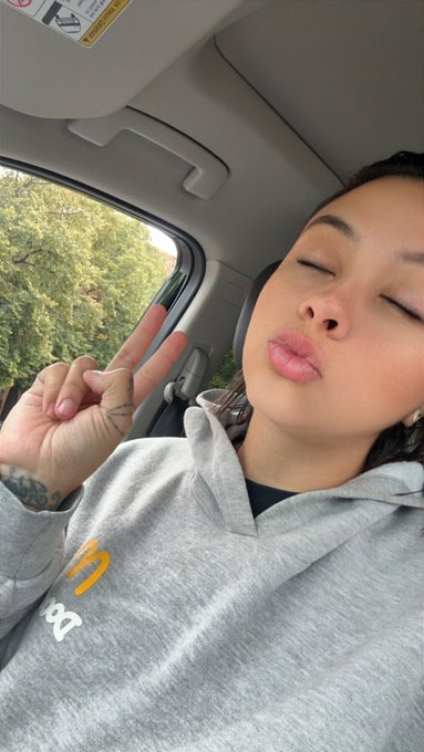A person wearing a gray hoodie makes a peace sign and puckers their lips for a selfie inside a car.