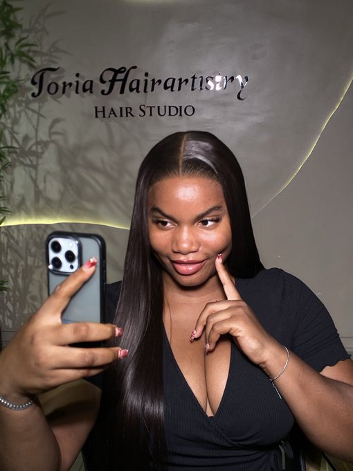 Mirror selfie of a woman at Toria Hairtistry Hair Studio showing off sleek, straight black hair, with pink nails and a black V-neck top.