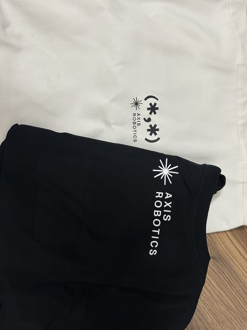 The image features a black t-shirt with white text that reads " Axis Robotics" along with three stars, possibly representing the company's logo or brand name. The t-shirt appears to be folded up, and there are two bags visible in the scene - one on top of the other.