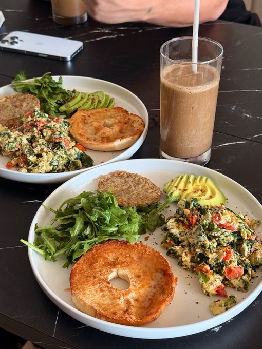 Two plates of food featuring bagels, scrambled eggs with vegetables, avocado, sausage patties, and arugula sit on a black table next to an iced coffee and an iPhone.