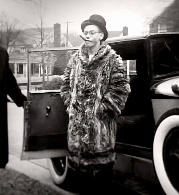 A vintage black-and-white photograph shows a young man wearing glasses, a top hat, and a heavy fur coat standing next to an open car door with a cigar in his mouth.