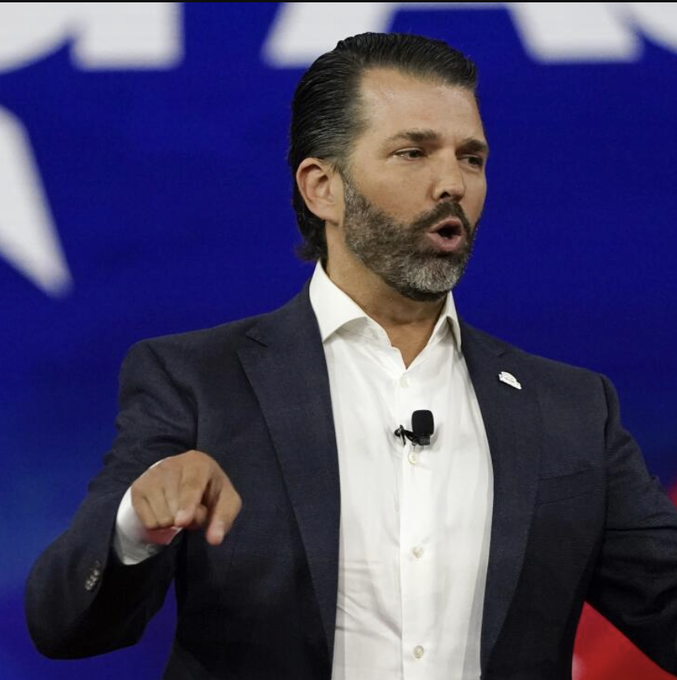 Donald Trump Jr., wearing a dark suit and white shirt, gestures emphatically while speaking at a podium against a blue backdrop—relevant as the post discusses World Liberty Financial ($WLFI), a crypto venture he is publicly associated with, delaying a token launch tied to a Trump resort due to Iran-related travel disruptions.