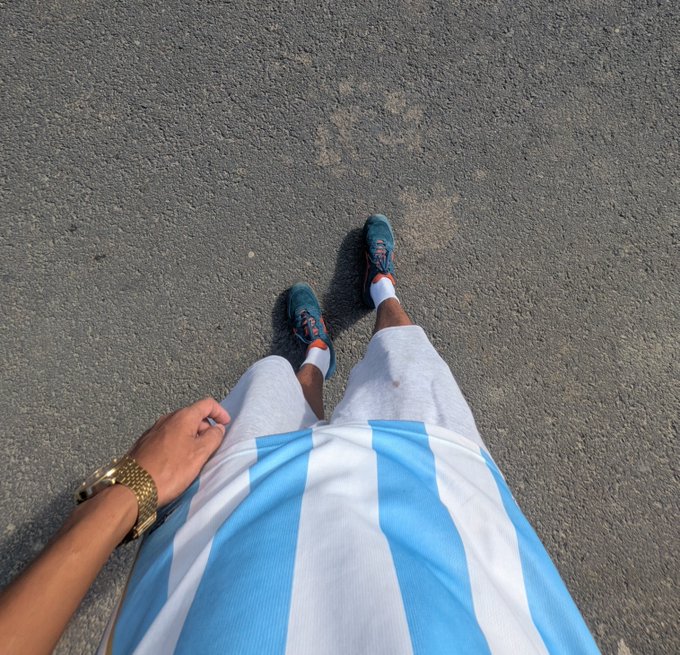 First-person view looking down at teal running shoes, white shorts, and a blue and white striped shirt on gray pavement, with a gold watch on the wrist.