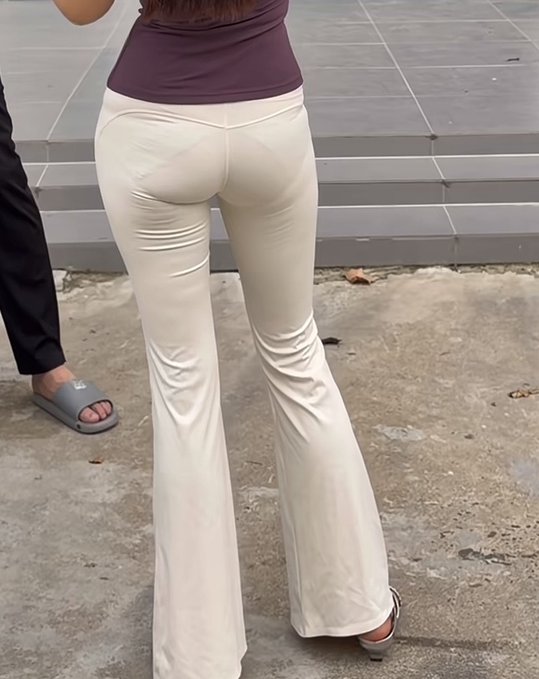 A woman poses outdoors in white flared leggings under a purple top, with the back seam and fabric folds resembling an open-crotch design. The caption jokes about how the suggestive pants leave her next move hard to predict.