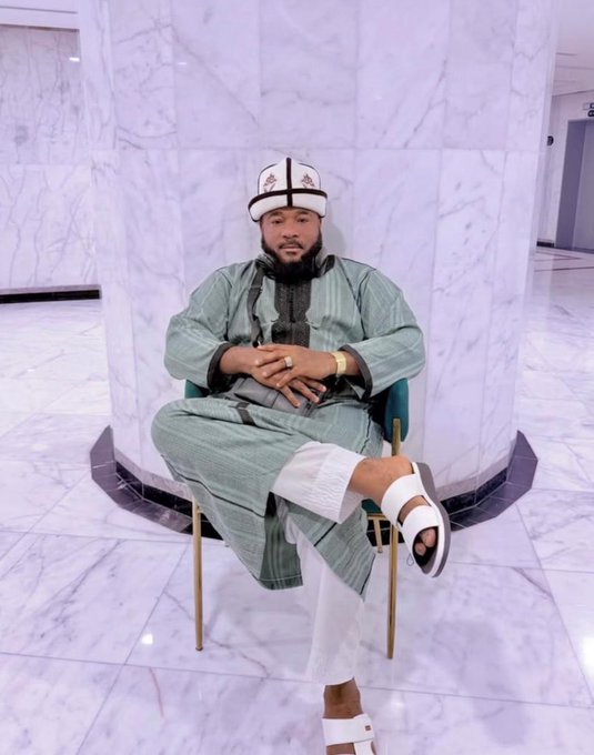 A man with a beard sits cross-legged in a chair, wearing a green traditional outfit, white pants, white sandals, and a white cap with decorative patterns, posed in front of a marble pillar.