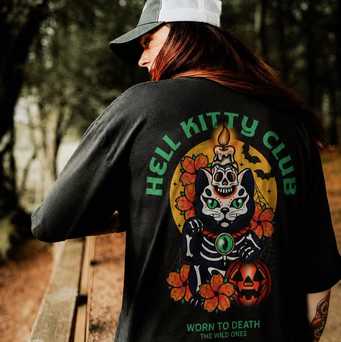 A person viewed from behind wearing a black baseball cap and a black t-shirt featuring a colorful graphic design. The shirt displays a skeleton cat with a candle on its head, a jack-o'-lantern, and the text 'HELL KITTY CLUB' at the top and 'WORN TO DEATH / THE WILD ONES' at the bottom.