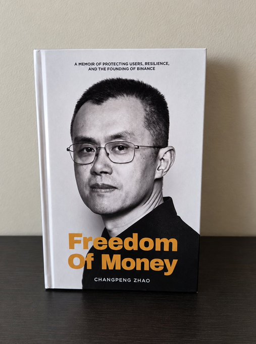 A book titled 'Freedom Of Money' by 'CHANGPENG ZHAO' stands upright on a dark surface, featuring a black-and-white portrait on the cover and the subtitle 'A MEMOIR OF PROTECTING USERS, RESILIENCE, AND THE FOUNDING OF BINANCE'.