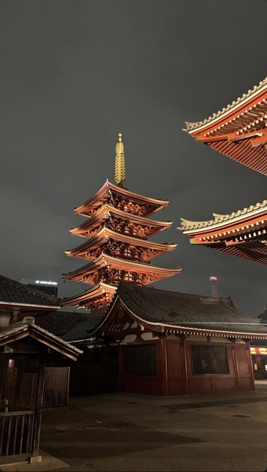 The image features a large, multi-tiered pagoda with gold accents standing tall against a dark sky at night. The pagoda appears to be lit up, drawing attention to its intricate design and architectural details. In the foreground of the photo, there is an old building that seems to be part of a larger structure or complex. A person can also be seen in the image, possibly admiring the view or passing by.