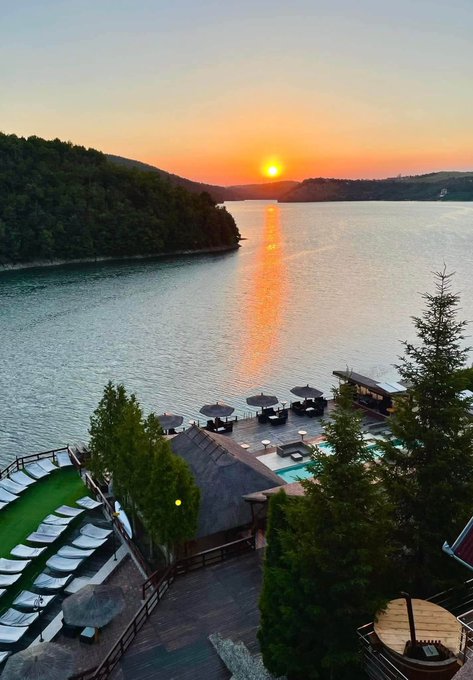 A scenic sunset view over a lake surrounded by forested hills, with a resort area in the foreground featuring a pool, lounge chairs, umbrellas, and a wooden deck.
