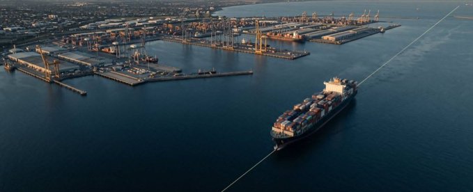An aerial view of a large cargo ship loaded with containers being towed through a harbor, with industrial port infrastructure and cranes visible in the background.