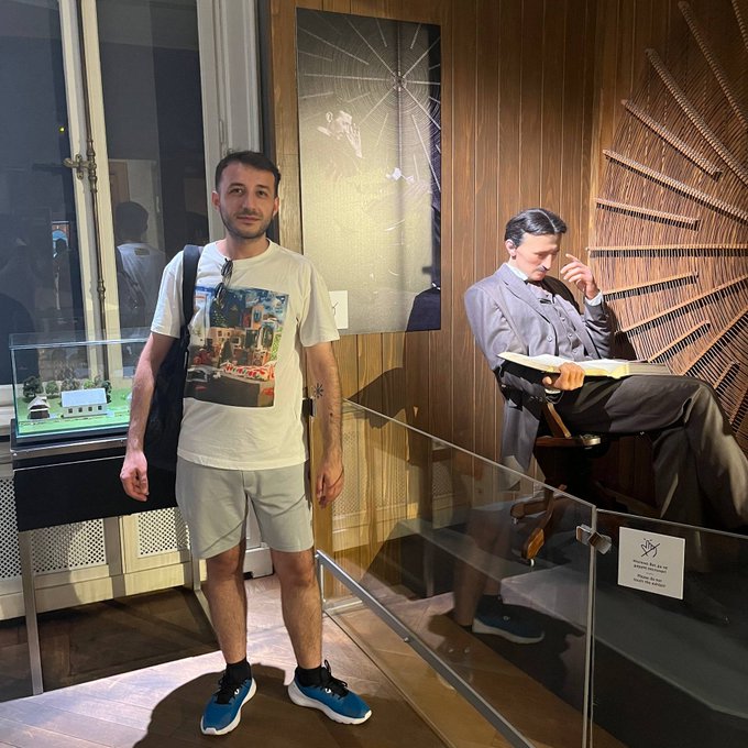 A man stands next to a glass display case containing a life-sized figure of a man in a gray suit sitting in a chair reading a book, with a plaque that appears to say 'Wernher' and 'Moonwalker'.