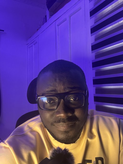A selfie of a man wearing glasses and a yellow sweatshirt, sitting in a room with purple lighting and striped window blinds.