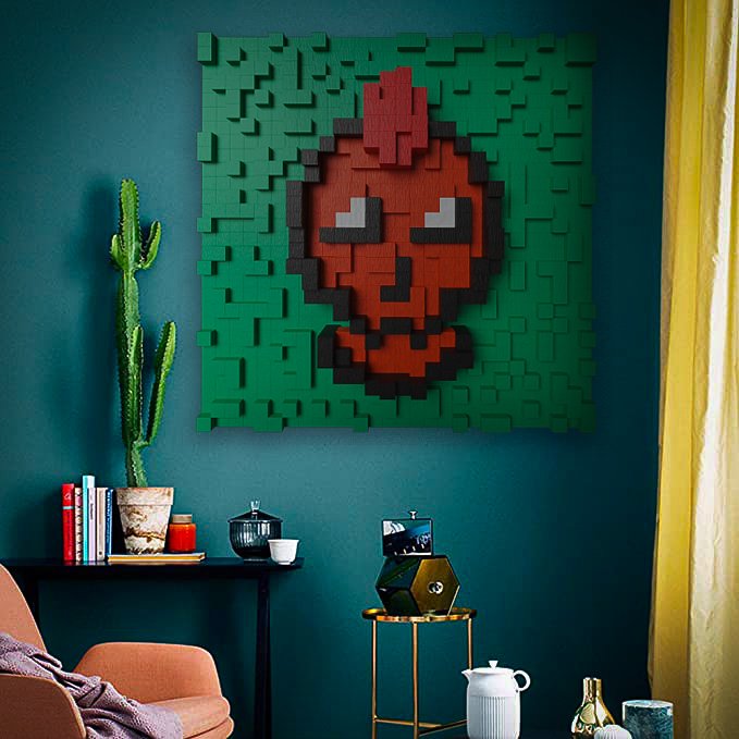 A LEGO-style pixel art wall decoration depicting a red Angry Birds character mounted on a teal wall, with a cactus plant, books, and a small side table visible in the room below.