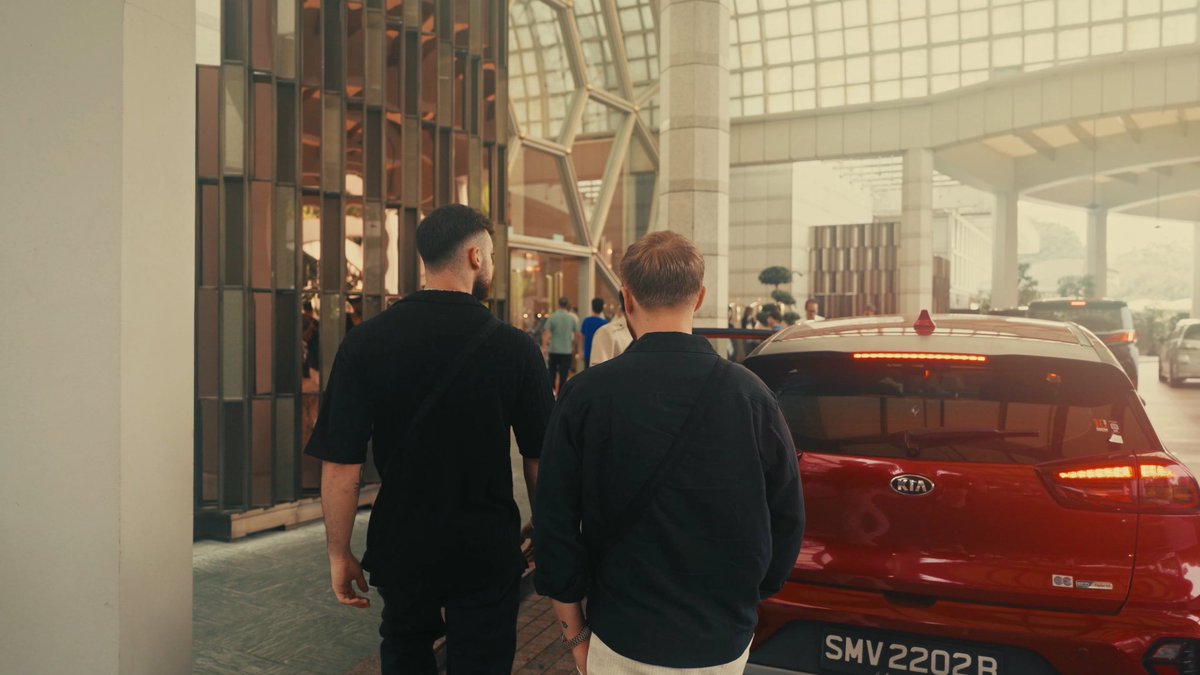 Two men walk toward a luxury building entrance beside a red Kia with the license plate "SMV 2202B," arriving in style at a high-end venue befitting the competitive, high-stakes nature of trading now framed as an esport on Solana.