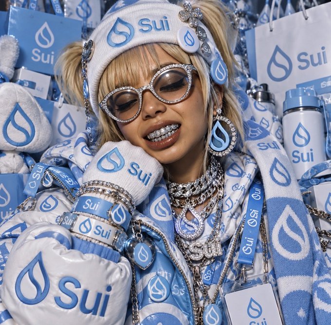 A blonde woman wearing rhinestone glasses, diamond grillz, and extensive 'Sui' branded apparel and accessories including a hat, gloves, boxing glove, and water bottle.