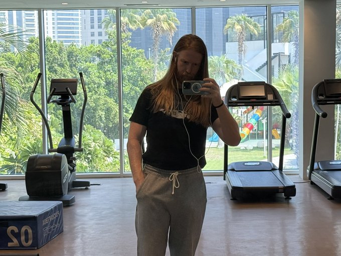 A long-haired man takes a mirror selfie in a gym with treadmills and an elliptical machine visible, with a box labeled "20 KG" and "JUST STRONG" in the foreground, while wearing earbuds and a "TS" shirt, humorously illustrating the post's punchline that he'll prioritize fitness gains over "Monad" crypto hype this cycle.