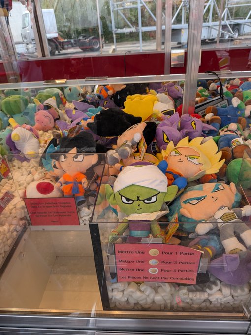 A claw machine filled with Dragon Ball plush toys, including recognizable characters like Goku and Piccolo, with a pricing sign in French indicating 1 token for 1 try, 2 tokens for 2 tries, and 5 tokens for 5 tries.