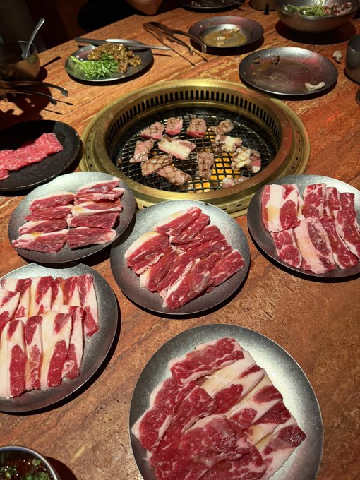 An overhead view of a Korean BBQ grill with multiple plates of raw, marbled beef slices arranged around the central cooking surface, along with side dishes.