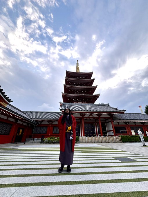 A person wearing a red traditional Japanese robe standing in the courtyard of a five-story pagoda temple, likely Senso-ji in Tokyo, under a dramatic cloudy sky.