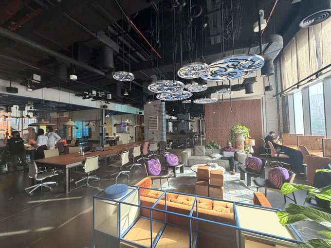 An interior view of a modern, industrial-style co-working space or lounge featuring distinctive circular ceiling lights with blue patterns, wooden communal tables, lounge seating areas, and exposed ductwork.
