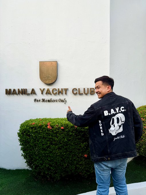 The image features a man wearing a black jacket with white text, standing next to a building that has a sign for "Manila Yacht Club". The man appears to be posing for a photo or attending an event at the club.