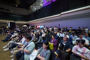 A large audience seated in rows at what appears to be a conference or tech event, with attendees looking toward a stage illuminated by purple lighting.