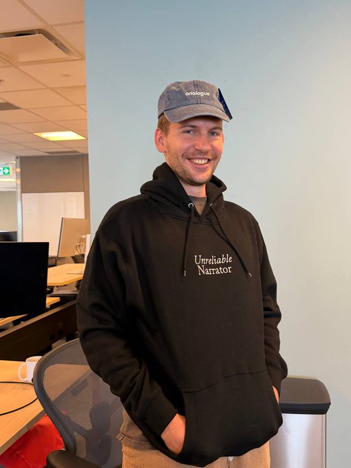 A man smiles in an office setting wearing a black hoodie with "Unreliable Narrator" printed on it and a grey cap reading "analogue." The self-aware "Unreliable Narrator" apparel aligns playfully with the post's complimentary caption about the subject.
