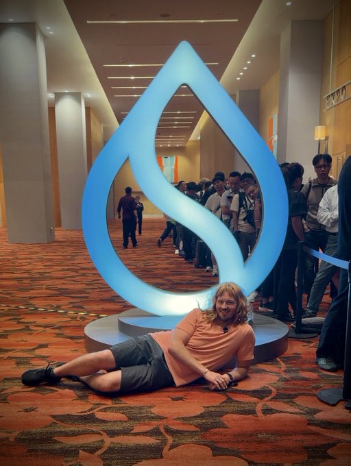 A person lounges on the floor in front of an illuminated Sui logo sculpture at what appears to be a conference or event, with attendees gathered nearby—visually reinforcing the confident declaration "$Sui will win."