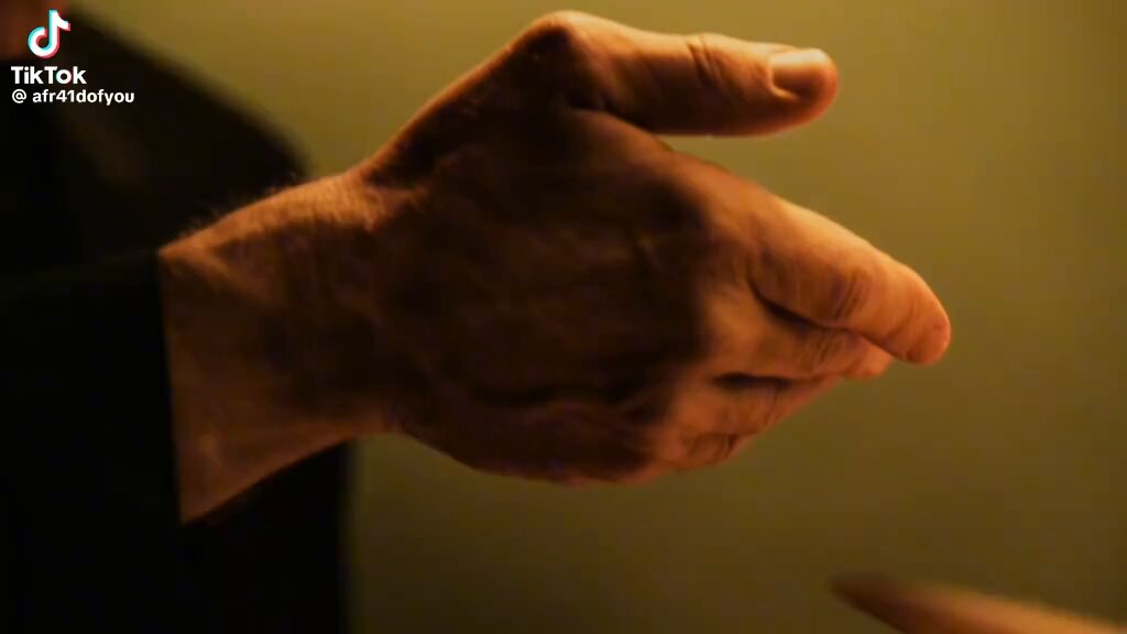 The image shows a close-up view of an older man's hand, with his palm facing upwards as if he is about to make a gesture or clap his hands together. The background appears to be a dark green color, which contrasts with the light brown hue of the man's hand and adds depth to the scene. There are no other objects in the image, and the focus is solely on the man's hand and its movement.