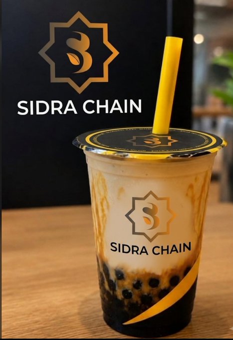 A plastic cup filled with creamy brown sugar bubble tea (likely tiger sugar boba) sits on a wooden counter in front of a black "SIDRA CHAIN" sign, featuring the brand's gold eight-pointed star logo. The drink's caramel-streaked appearance complements the post's playful hint about community members enjoying ice cream this month, suggesting a fun seasonal treat alongside the #SidraChain #SDA #Blockchain branding.