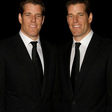 Two men in matching black suits and ties stand smiling side by side against a dark background, their nearly identical appearance visually reflecting the Winklevoss twins referenced in the post about Gemini's ongoing financial struggles.