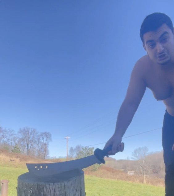 A shirtless man outdoors under a blue sky holds a large knife resting on a wooden stump.