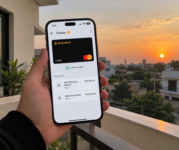 Hand holding a smartphone displaying a 'Poison' cryptocurrency wallet app by Binance showing card details ending in 2754 and transaction history, set against a sunset view over a cityscape.