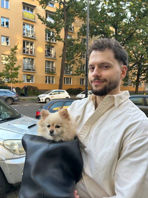 A bearded man in a cream-colored shirt holds a small fluffy Pomeranian dog in a black bag on a city street.