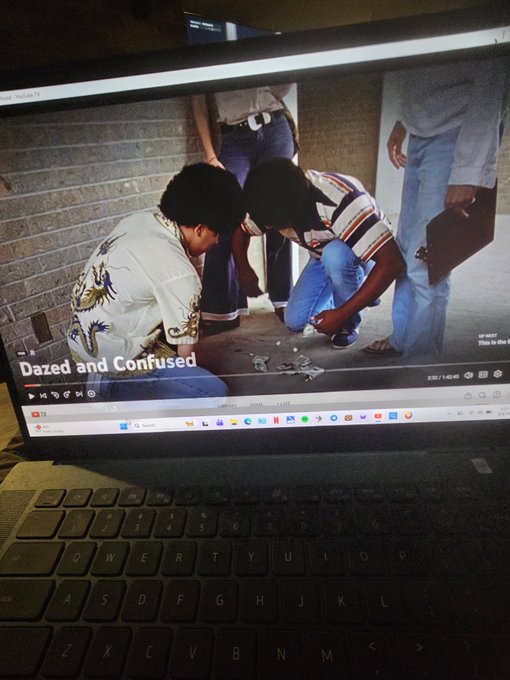 A laptop screen displaying the video 'Dazed and Confused' showing two men crouched on the ground examining something, with the laptop's black keyboard visible in the foreground.