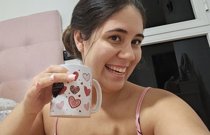 A smiling young woman holds a white mug decorated with red and pink hearts, posed in front of a pink tufted headboard.