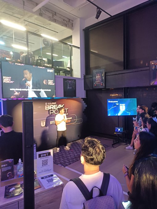 A dimly lit event space where an audience watches a presentation, with a screen displaying 'POINT FORM BYT' and 'BYB'T', and another screen showing 'Welcome to BYT'.