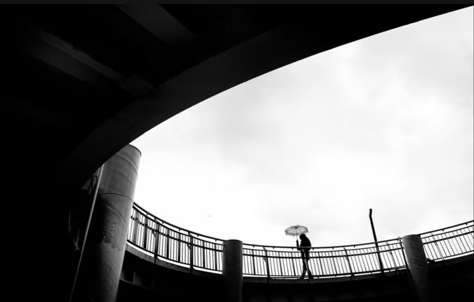 A black and white photograph taken from below showing a person with an umbrella walking along a curved elevated walkway or bridge against a cloudy sky.
