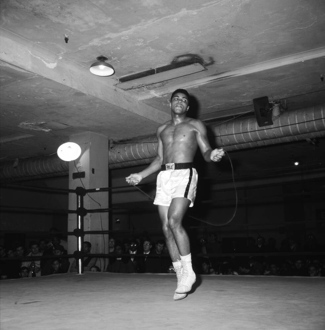 A black and white historical photograph of a shirtless Black boxer wearing white shorts and Everlast boots, skipping rope in a boxing ring before a crowd.