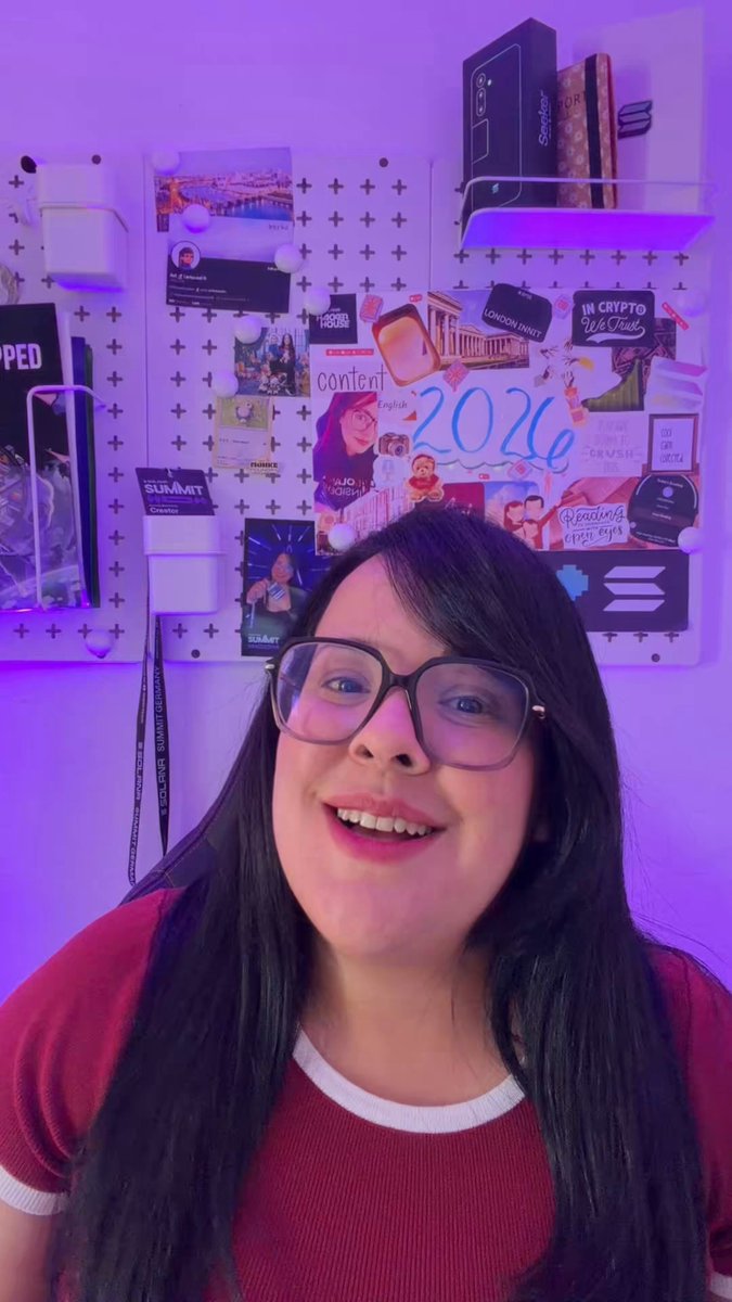 The image features a young woman with long black hair wearing glasses and a red shirt, smiling at the camera while posing for a photo. The background of the image includes various posters and pictures, suggesting that she might be attending an event or gathering related to the Solana Summit Germany.