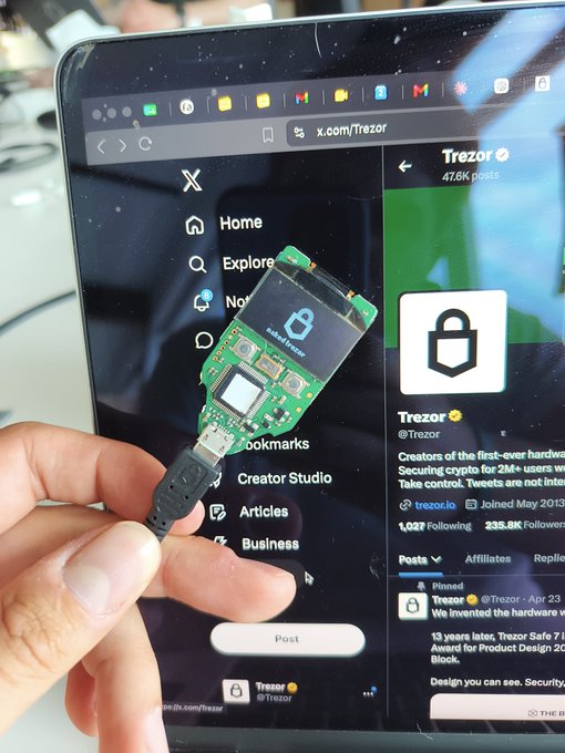 The image shows a person holding up a small green device, which appears to be a Trezor, connected by a black cord or USB cable. The device has a screen that displays a website called "trezor.com". This suggests that the person might be interested in learning more about Trezor devices and their features, as well as possibly considering purchasing one for themselves.