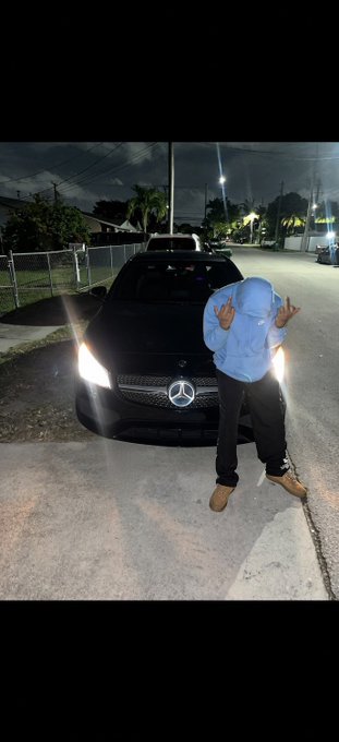 A person in a light blue hoodie and Timberland boots poses in front of a black Mercedes-Benz at night, making hand gestures toward the camera.
