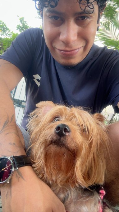 A close-up selfie shows a young man with curly hair and arm tattoos smiling while holding a small brown Yorkshire Terrier dog.