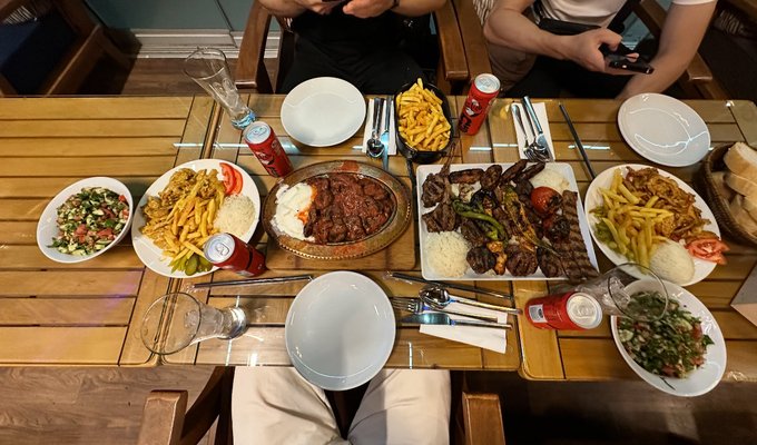 A wooden table is set with a feast featuring various meat dishes, including grilled kebabs and stews served over rice, alongside plates of french fries, fresh salads, tomatoes, and bread, accompanied by cans of Coca-Cola and empty place settings. Two people are partially visible seated at the table, with one holding a smartphone.